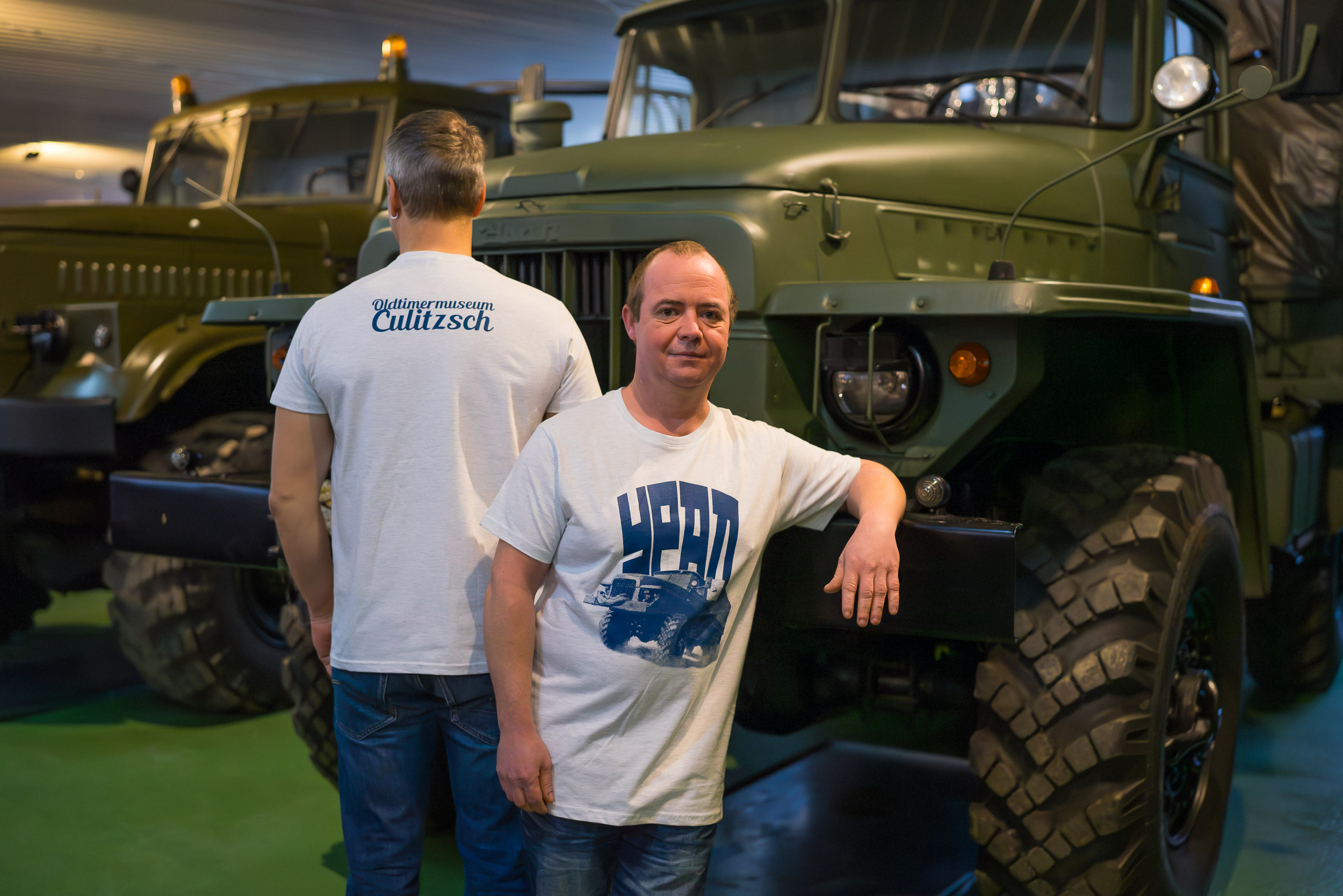 zwei Männer vor Ural-Auto im Museum, einer mit Gesicht zur Kamera, anderer mit Rücken zur Kamera. Beide tragen grau meliertes T-Shirt mit dunkelblauem Ural-Print und dunkelblauer Aufschrift URAL in kyrillischen Buchstaben.
