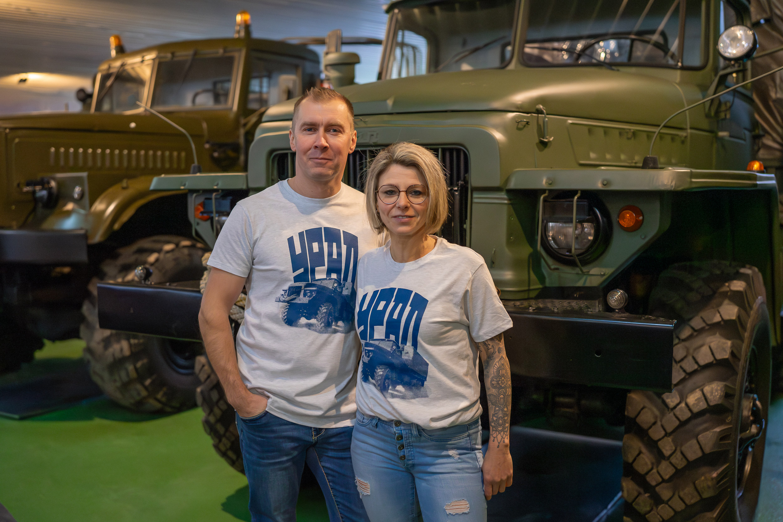 Frau und Mann vor Ural im Museum, beide tragen grau meliertes T-Shirt mit dunkelblauem Ural-Print und dunkelblauer Aufschrift URAL in kyrillisch