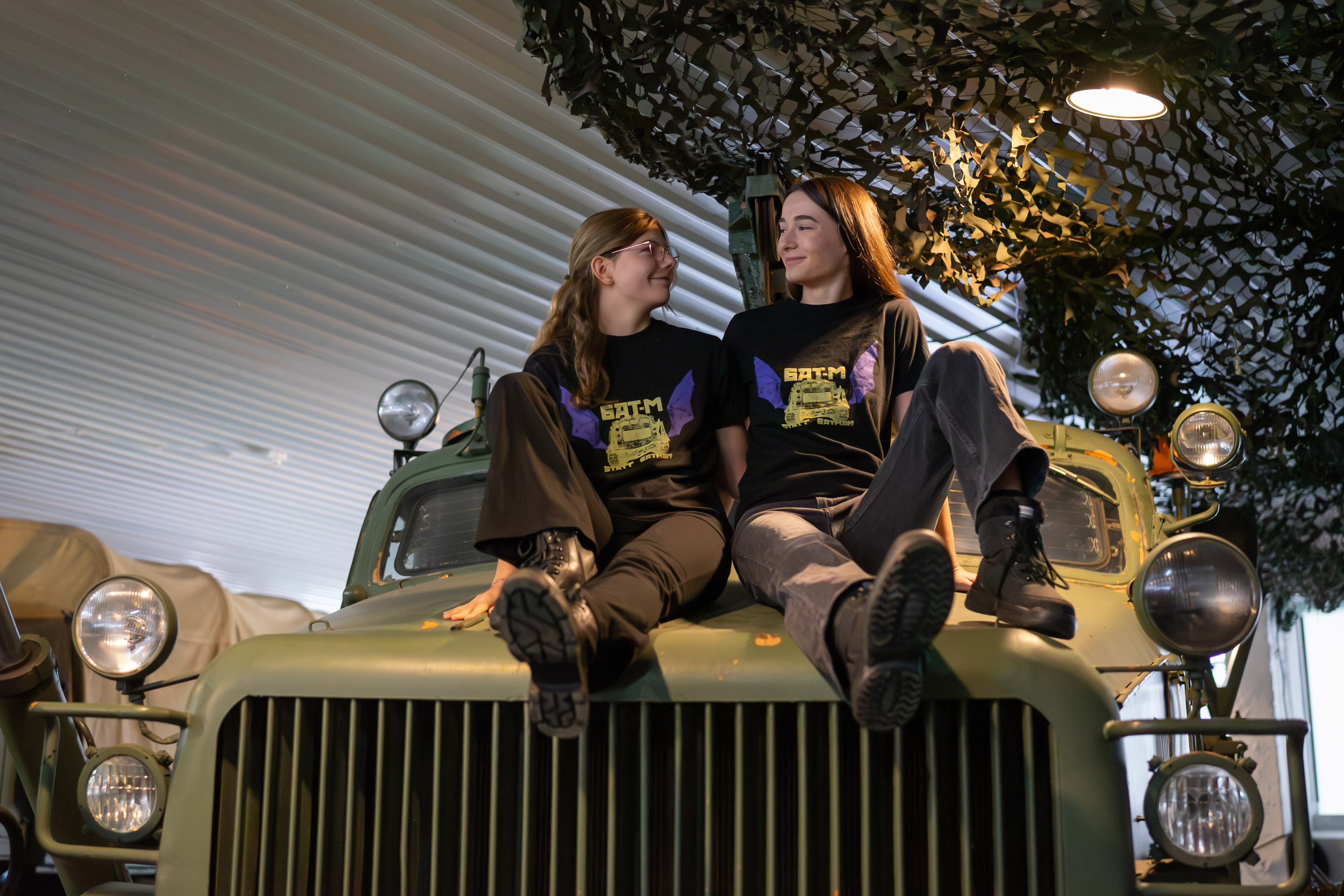zwei junge Frauen auf Motorhaube der BAT-M, beide tragen schwarzes T-Shirt mit gelber Aufschrift BAT-M statt Batman und lila Print BAT-M-Raupe mit Fledermausflügeln.